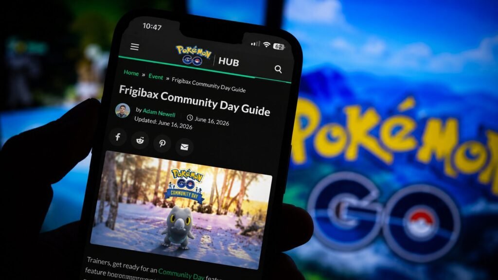 A hand holds a smartphone displaying a Pokémon GO Hub article titled Frigibax Community Day Guide. The screen shows a featured image of Frigibax in a snowy setting, while a blurred Pokémon GO logo appears in the background.